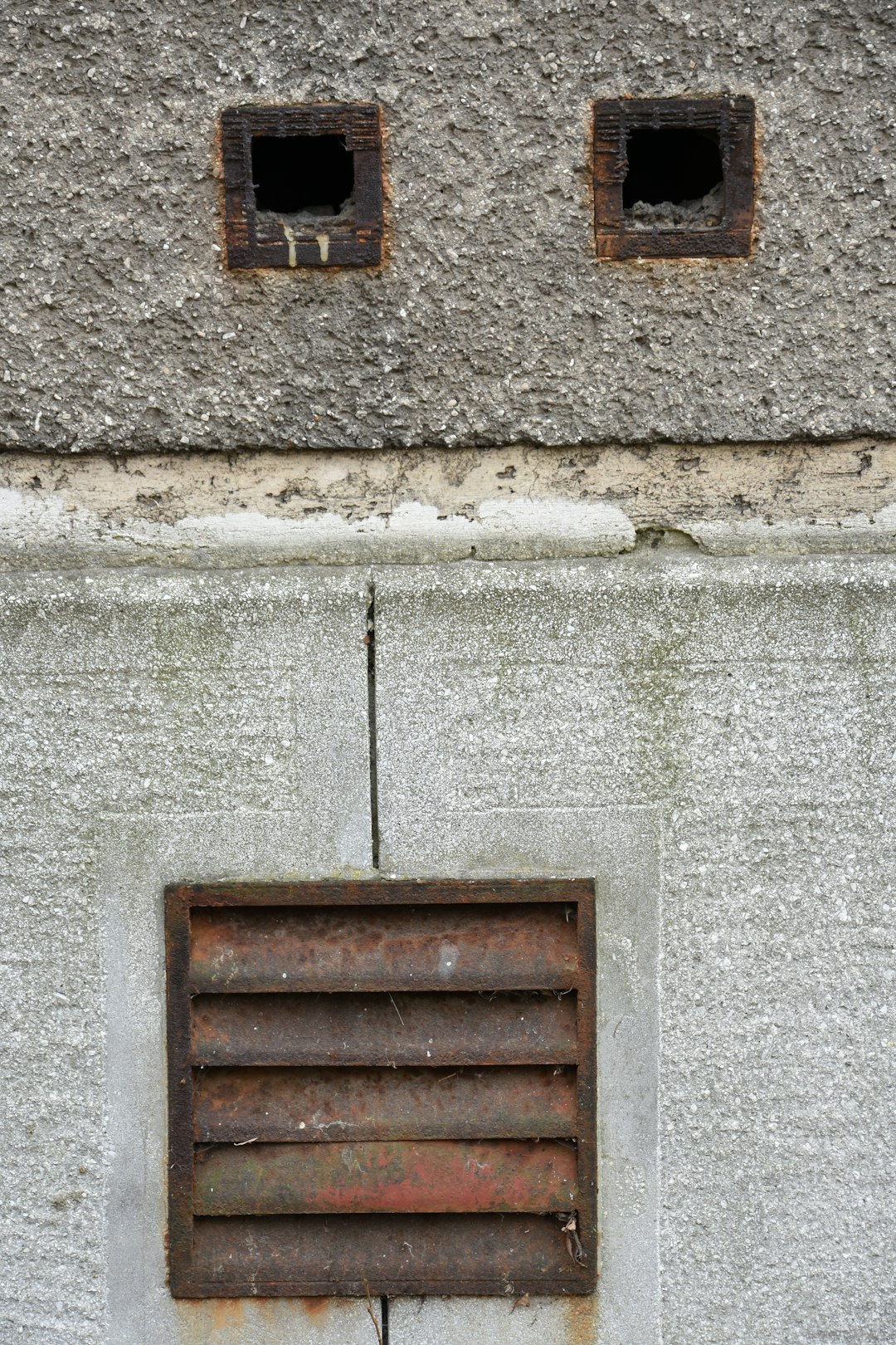 Two square openings above a rusty vent on wall