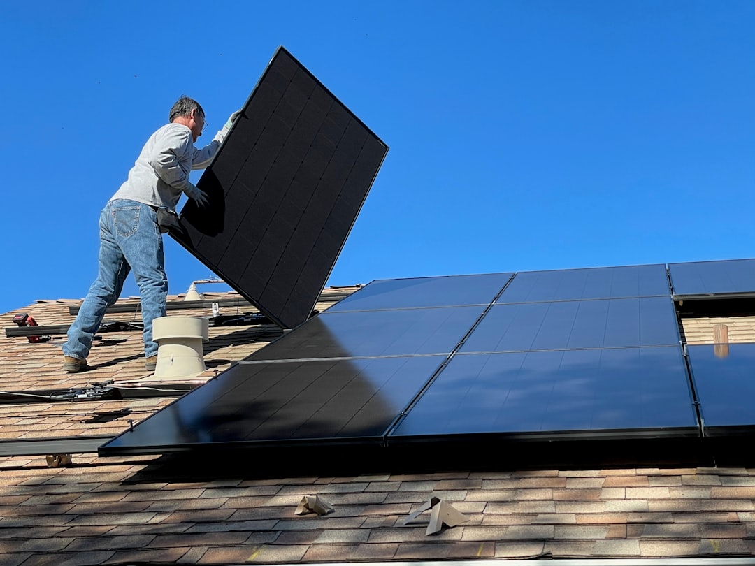 A person installing a black solar panel on a shingled roof under blue sky